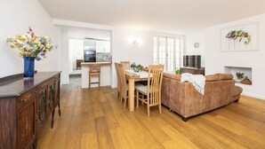 A modern living room with a brown sofa, wooden dining table, and chairs. There is a vase with flowers on a wooden cabinet. A kitchen is visible in the background. - Stunning Period Character Courtyard Coastal Flat - Pass the Keys (Hove)