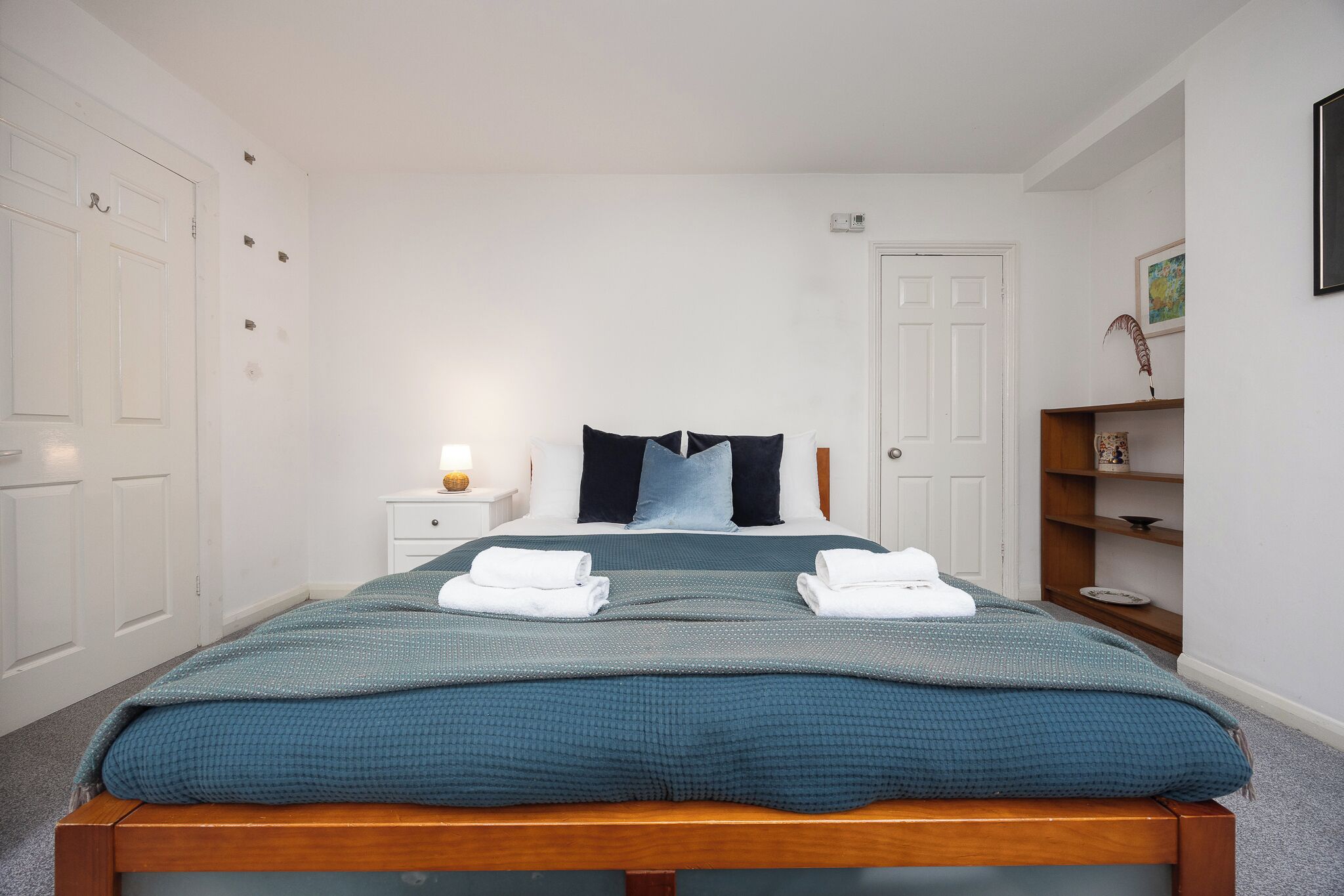 Un lit bien fait, avec une couette turquoise, une table de chevet avec une lampe et une étagère ornée d’objets décoratifs.