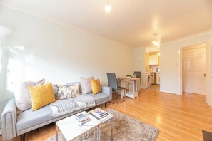 Ein modernes Wohnzimmer mit einem grauen Sofa, einem Couchtisch mit Büchern, einem Schreibtisch und einer Küche im Hintergrund.