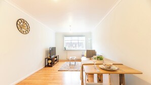 Un salon moderne avec un plancher en bois, une table à manger agrémentée d’un vase et d’un saladier, une horloge au mur, une fenêtre avec des stores et un radiateur.