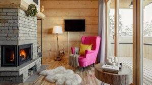 Une pièce chaleureuse avec une cheminée, un fauteuil rose, un téléviseur et une vue sur l’extérieur.