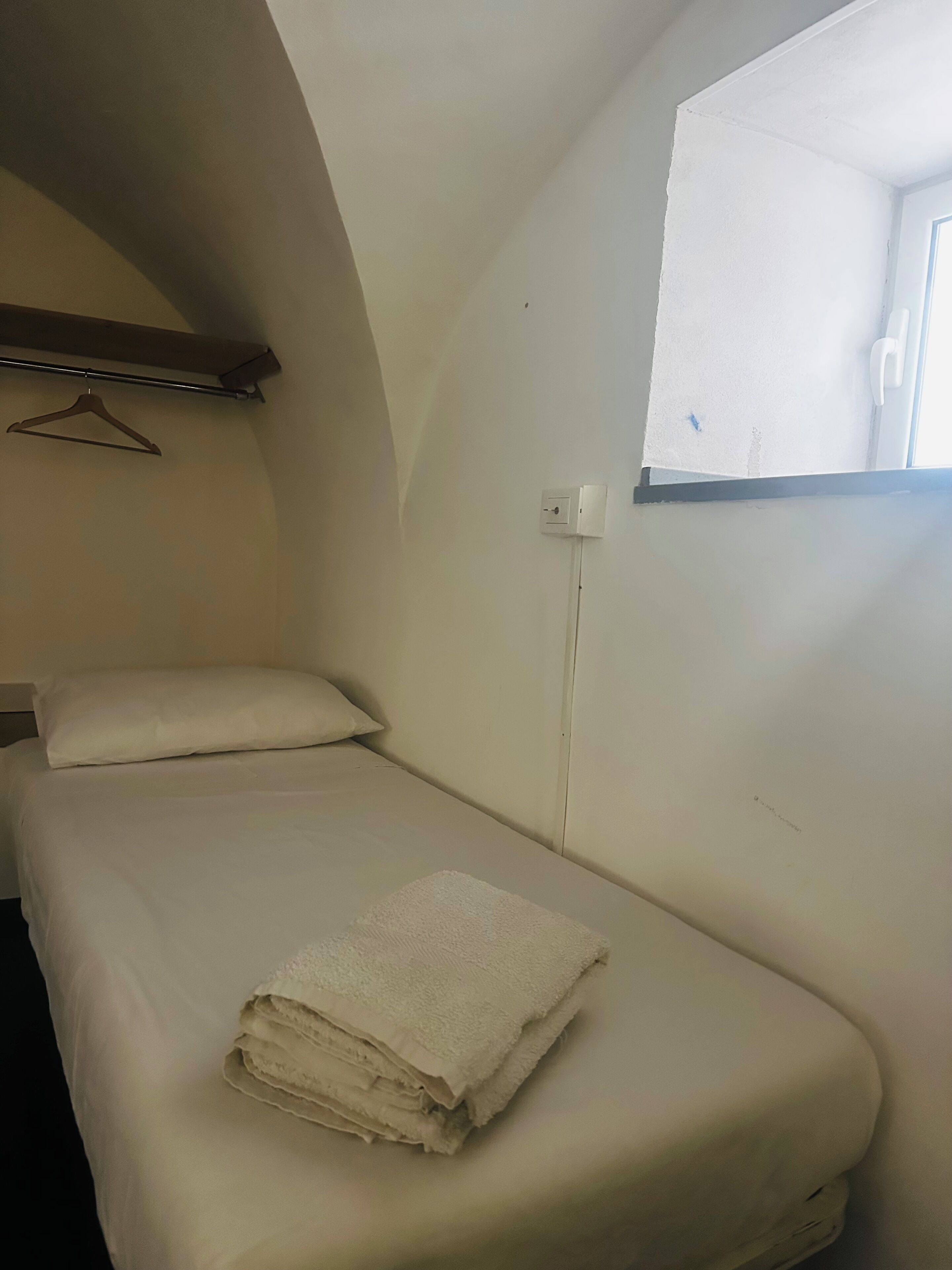 A small, single-bed room with a window, a hanging coat rack, and a power outlet on the wall.