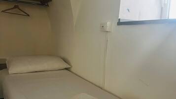A small, single-bed room with a window, a hanging coat rack, and a power outlet on the wall.