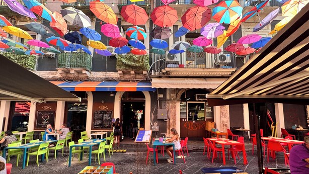 Ein Straßencafé mit bunten Sonnenschirmen, grünen und roten Stühlen und Tischen mit Speisekarten.