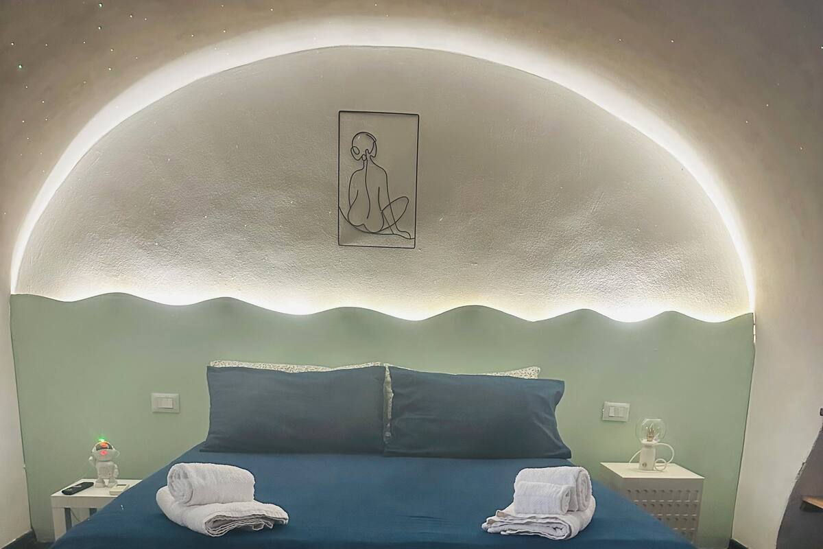 Une chambre à coucher avec un plafond incurvé, un lit avec une literie bleue, deux oreillers, un tapis moelleux et un tableau accroché au mur.