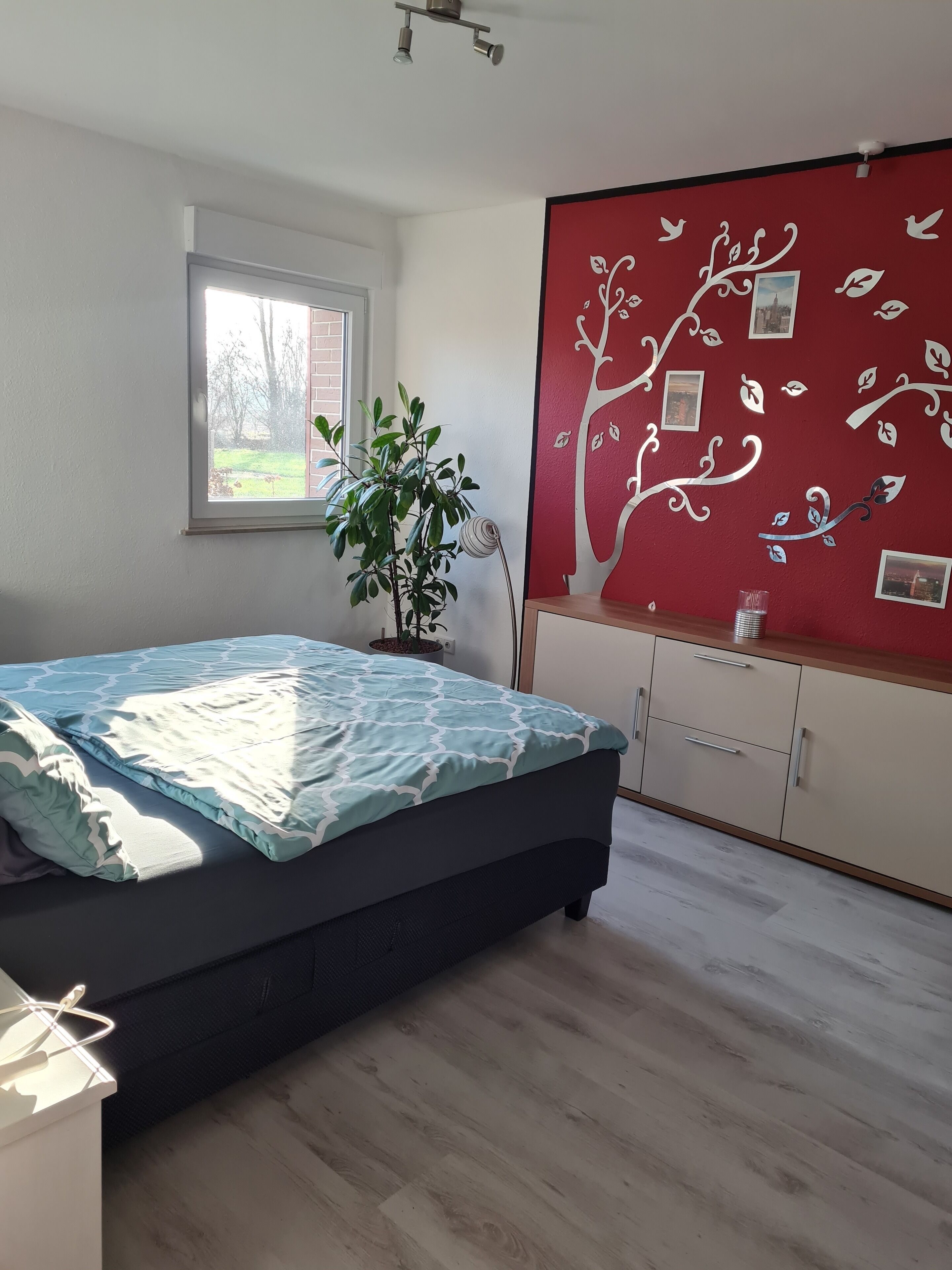 Une chambre à coucher avec un lit, une plante en pot, un mur rouge orné d’un motif d’arbre et une fenêtre.