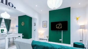 A modern living room with a green accent wall, a flat-screen TV mounted on the wall, a white sofa, and a dining area with a white table and chairs. - Modern 2 - Bed House Near Manchester City Centre - Sleeps 6 (Salford)