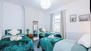 A hotel room with two beds, each with green bedding and white pillows, a mirror, a nightstand with a lamp, and a window with curtains. - Modern 2 - Bed House Near Manchester City Centre - Sleeps 6 (Salford)