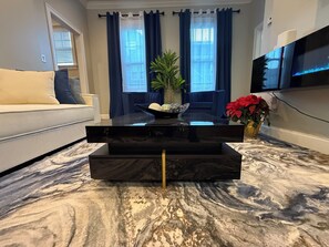 Un salon moderne avec un meuble de rangement noir pour les appareils électroniques, un sol à motifs de marbre, un canapé blanc, une table à manger avec des rideaux bleus et un téléviseur fixé au mur.