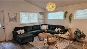 Ein Wohnzimmer mit einem dunkelgrünen Ecksofa, einem runden Couchtisch und Korbstühlen. Zwei Fenster mit Jalousien, eine Hängeleuchte und Wanddekorationen sind vorhanden.