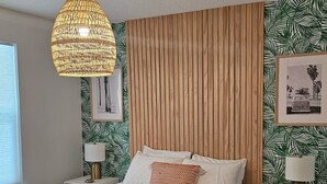 Ein modernes Schlafzimmer mit einem Holzbettgestell, einer gewobenen Pendelleuchte und Tapete mit tropischen Blattmotiven.