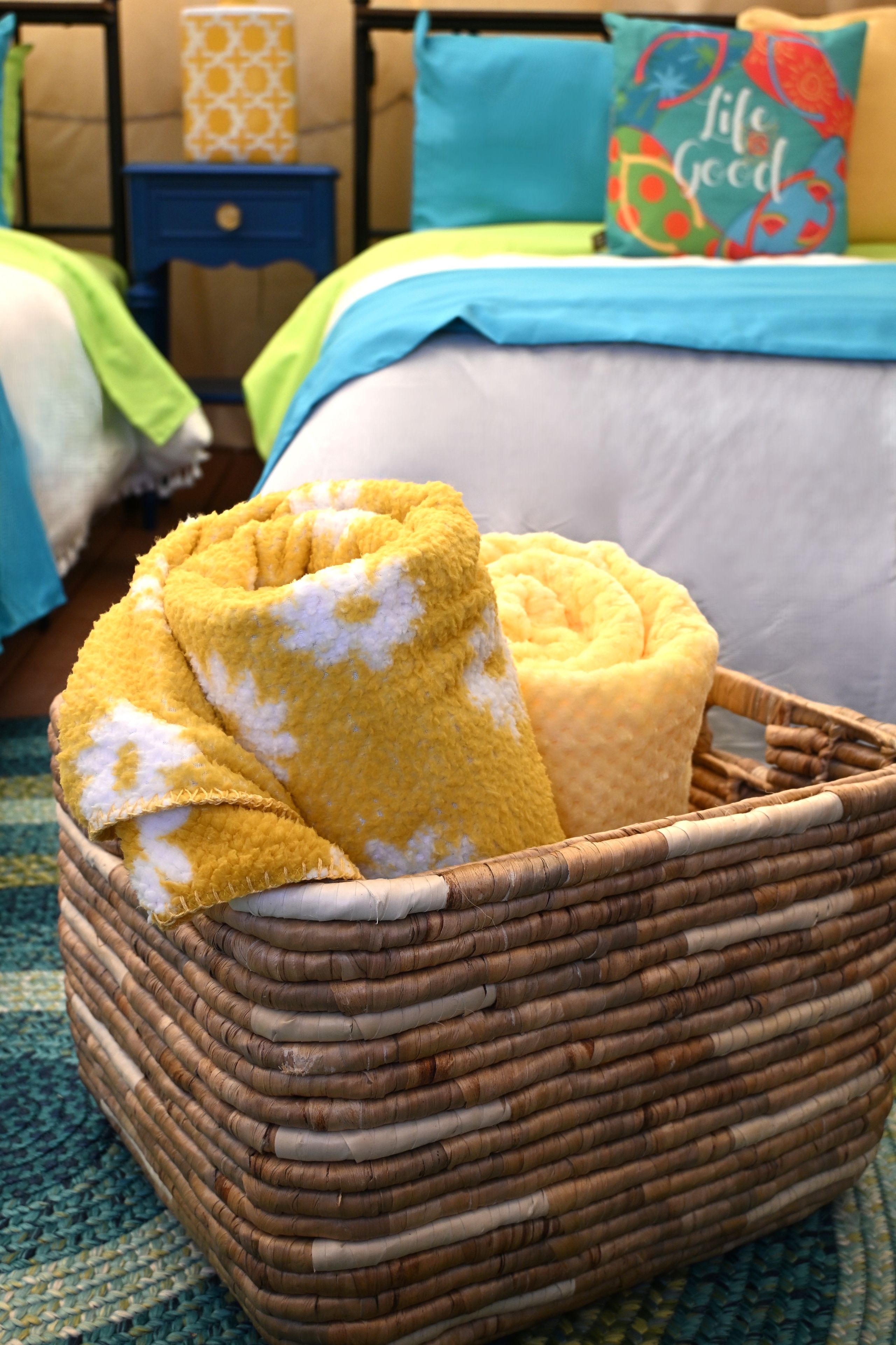 A bed with a blue and white bedspread, a blue nightstand with a lamp, and a colorful pillow with the text 'life is good.'