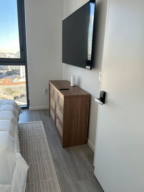 Ein Schlafzimmer mit einem Bett, einer Kommode, einem an der Wand montierten Fernseher und einem Fenster mit Blick auf die Stadt.