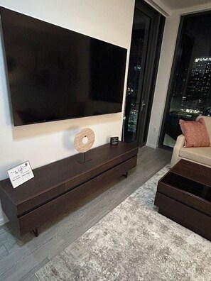 Ein modernes Wohnzimmer mit einem großen Flachbildfernseher an der Wand, einer niedrigen Unterhaltungsecke aus dunklem Holz und Blick auf die Stadt durch ein großes Fenster.
