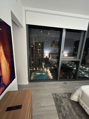 Un dormitorio con una ventana grande, una cama, un televisor y un control remoto sobre una mesa de madera.