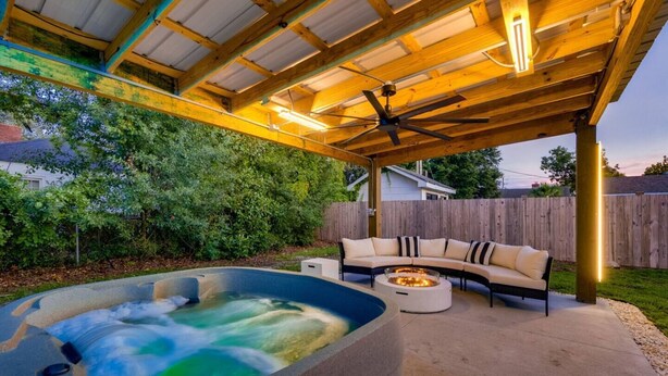 Een terras buiten met een jacuzzi, een ronde tafel met vuurkorf en een bank onder een houten pergola.