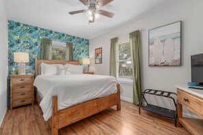 A bedroom with a wooden bed, a ceiling fan, a nightstand with a lamp, a desk with a chair, and a window with curtains. - Old Oak Oasis|Hot Tub·Golf Cart·Fire Pit·MHC (Morehead City)
