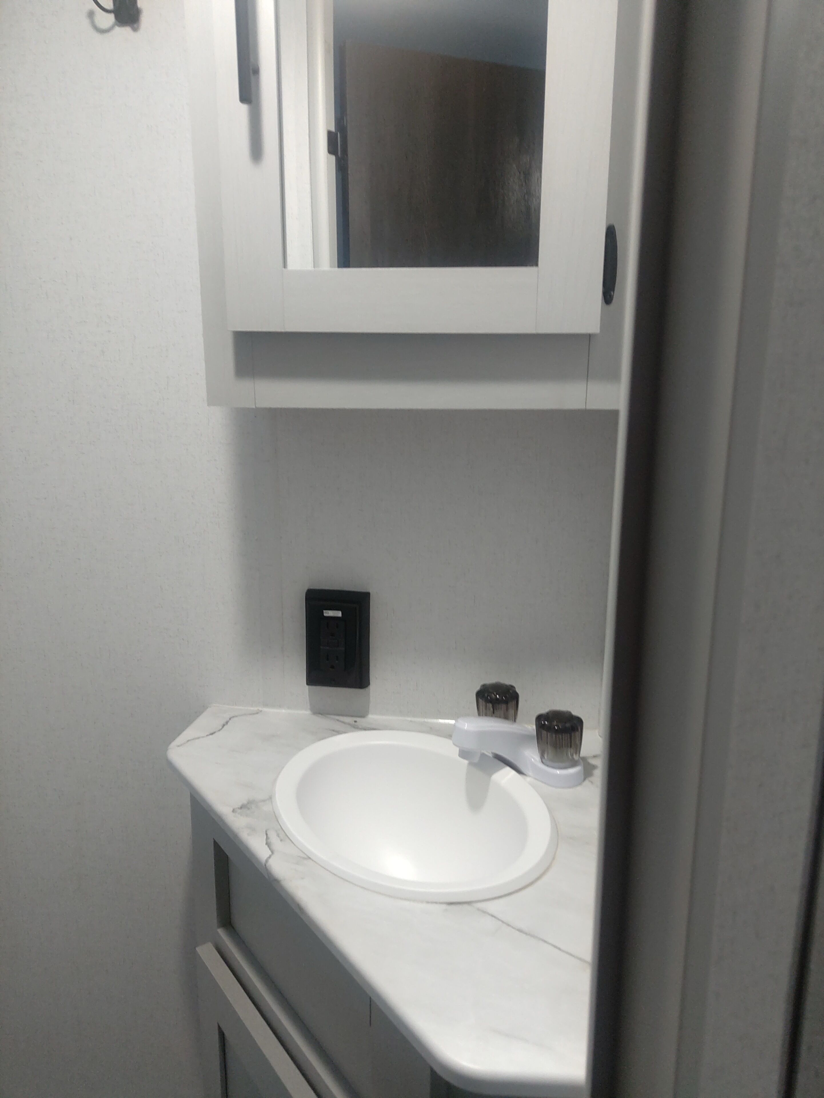 Un baño con un lavamanos blanco, un espejo encima y un pequeño enchufe negro en la pared.