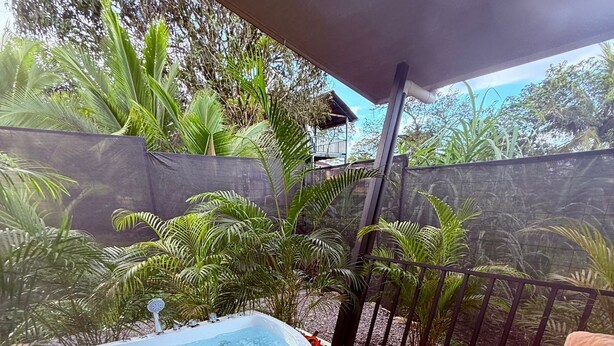 Ofurô numa varanda de madeira com vista para plantas tropicais e uma casa ao fundo.