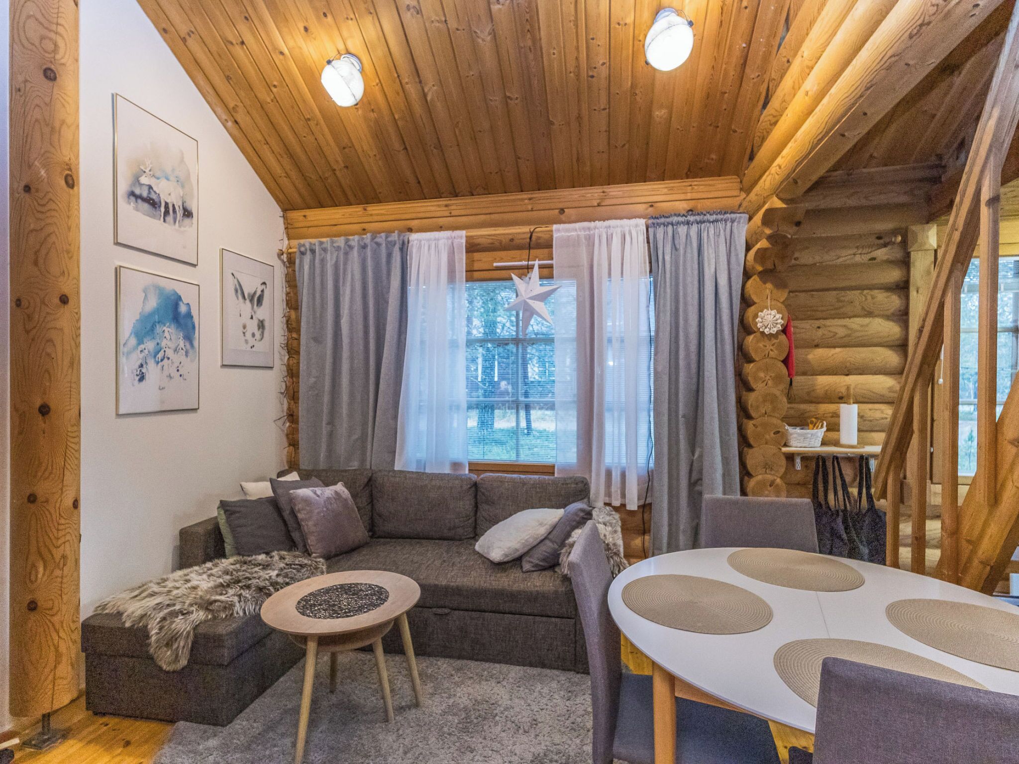 A cozy log cabin interior with a wooden ceiling, a gray sofa, a small round table, and a dining table with placemats.