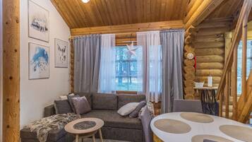 A cozy log cabin interior with a wooden ceiling, a gray sofa, a small round table, and a dining table with placemats.