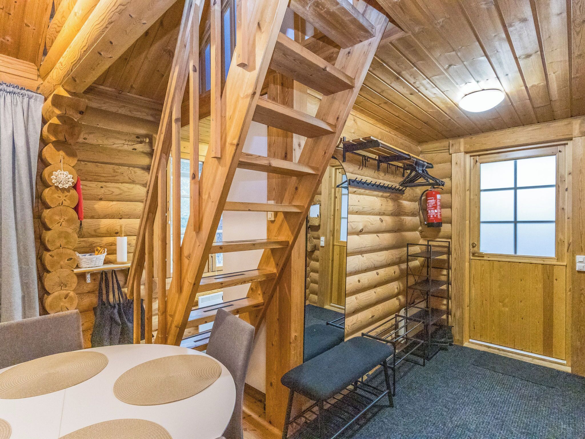 A log cabin interior with a wooden staircase, a dining table, and a fire extinguisher mounted on the wall.