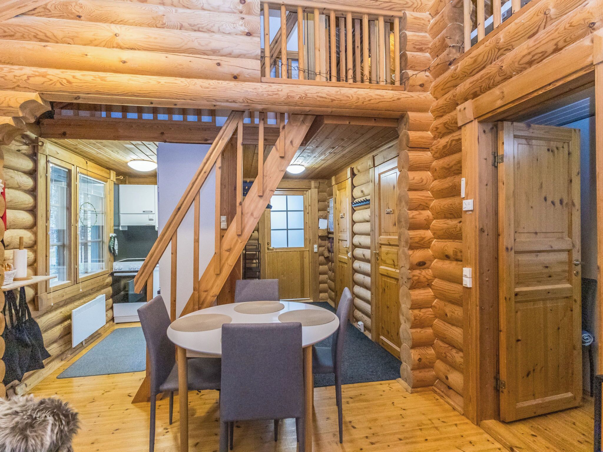 A log cabin interior with a dining area, a staircase, and a kitchen visible through an open door.