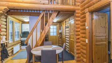 A log cabin interior with a dining area, a staircase, and a kitchen visible through an open door.