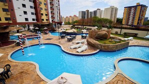 A swimming pool with multiple sections, lounge chairs, umbrellas, and a bridge. - Comfort and leisure, apartment in the Golden Dolphin Express hotel, fun for the family. (Caldas Novas)
