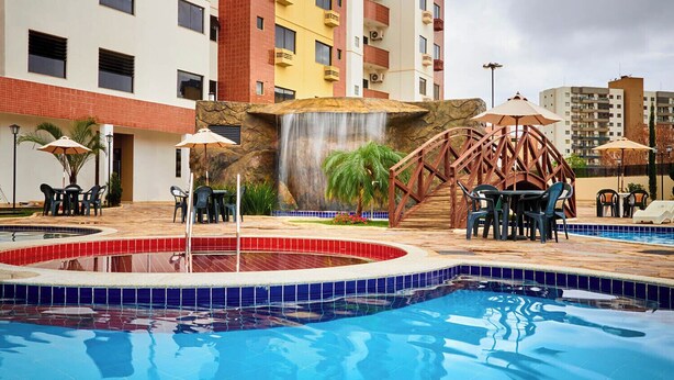 A hotel pool area with a waterfall feature, multiple seating areas, and a surrounding pool with tiled edges. - Comfort and leisure, apartment in the Golden Dolphin Express hotel, fun for the family. (Caldas Novas)