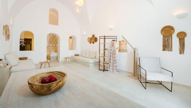 A spacious, well-lit room with a high arched ceiling, a wooden dining table with a bowl of fruit, a white sofa, a chair, and a small table with a lamp. - Iriana Apartments (Santorini)
