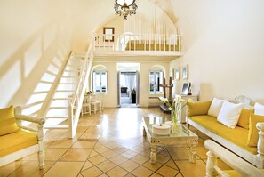 A spacious living area with a high ceiling, a staircase, and a view to the outside. - Enigma Apartment (Santorini)