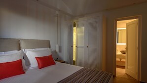 A hotel room with a large bed, white bedding, and a striped blanket. There is a wooden nightstand with a lamp and a red pillow. The room has white walls and a wooden floor. - Sea Dream (Santorini)