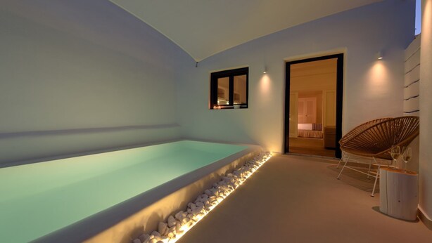 A modern indoor swimming pool with a pebble-lined edge, a wicker chair, and a small table with glasses. - Sea Dream (Santorini)