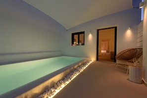 Ein modernes Hallenbad mit einem kiesverkleideten Beckenrand, ein Korbstuhl und ein kleiner Tisch mit Gläsern.