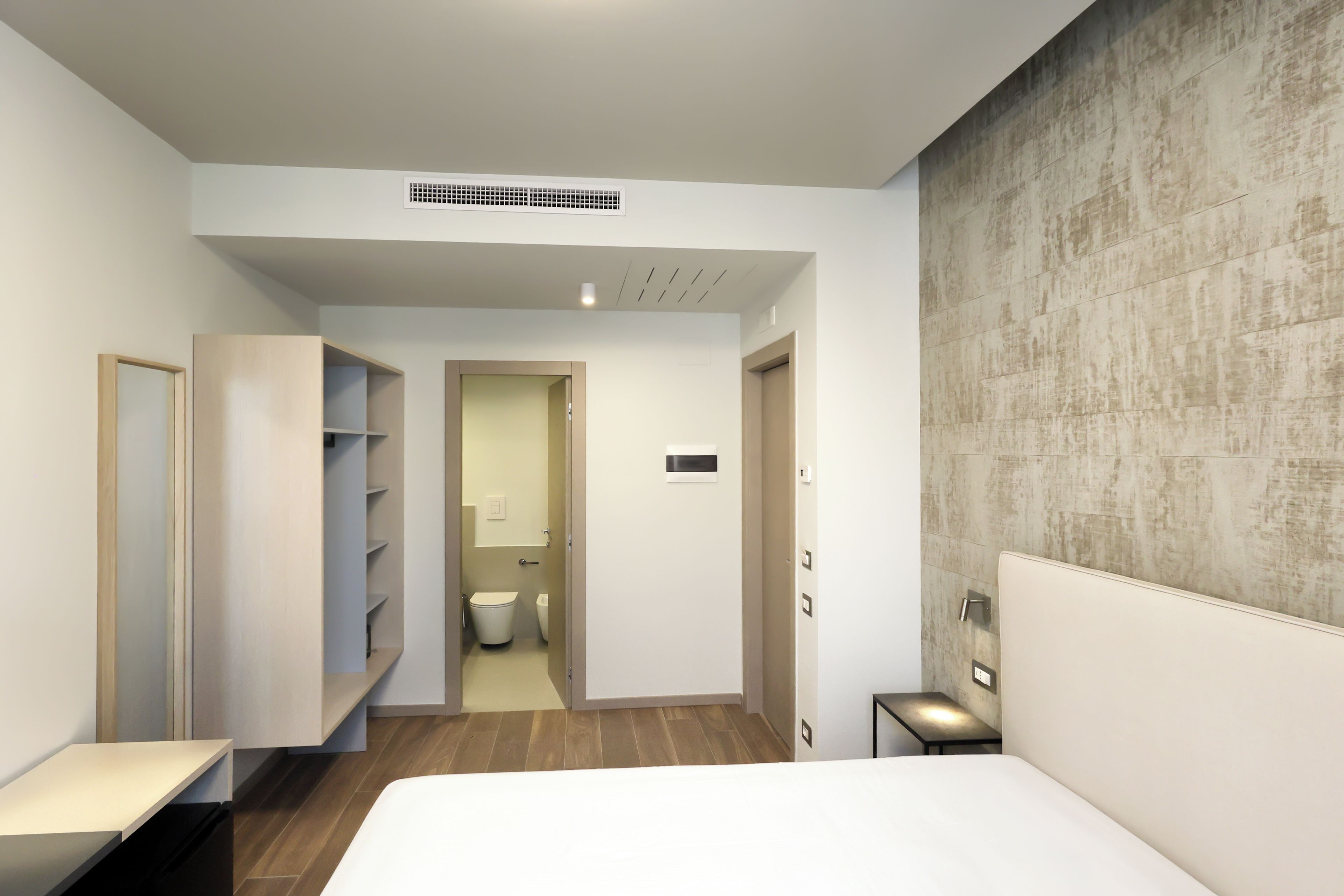Habitació d'hotel moderna amb un llit, una taula de nit, un armari, un bany amb vàter i un lavabo, i una paret amb relleu.