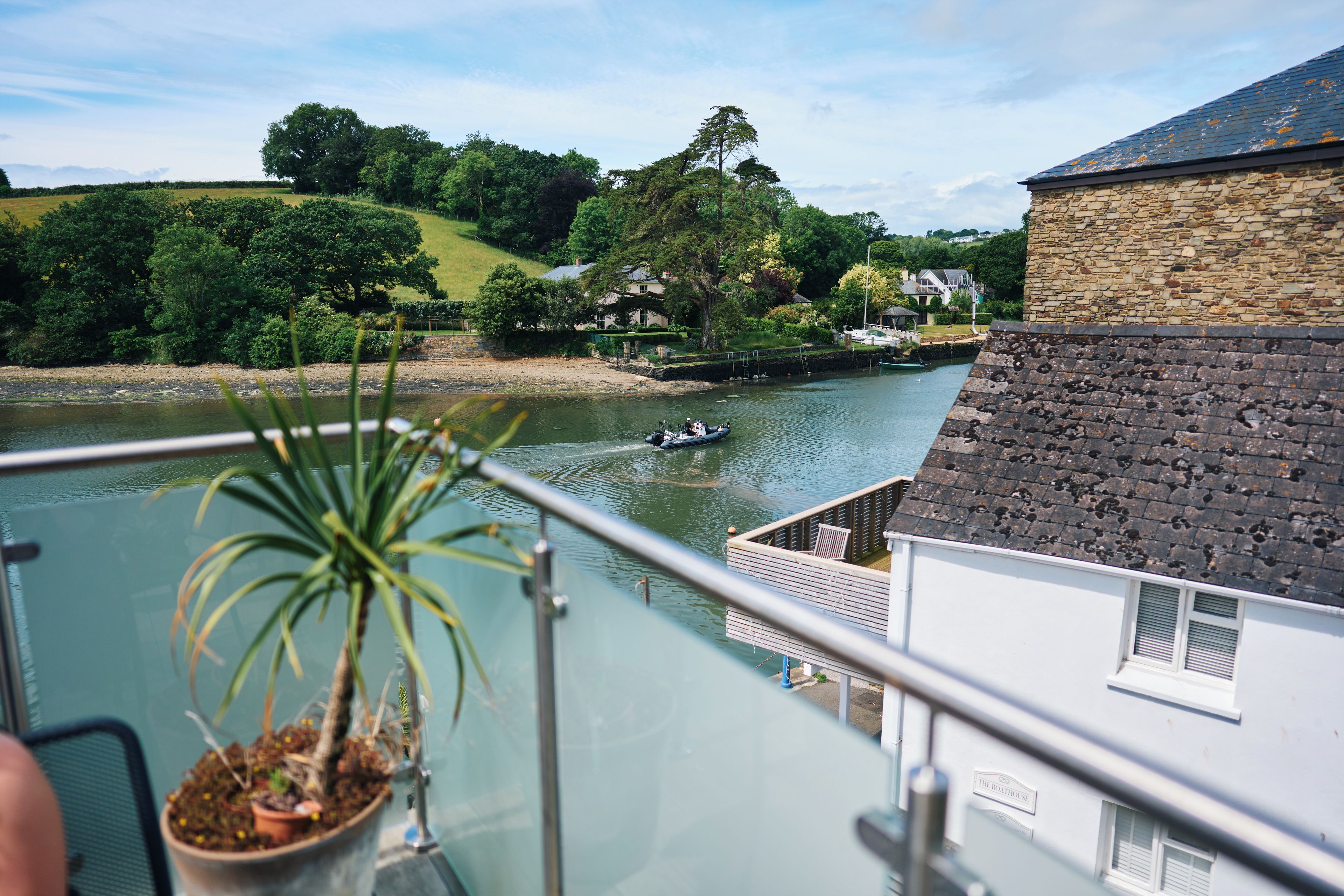 A riverside view from a balcony with a potted plant, a boat on the water, and a stone building in the background.