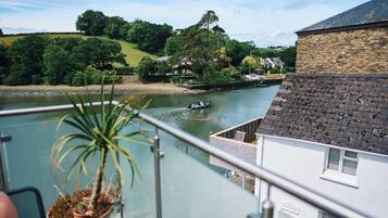 A riverside view from a balcony with a potted plant, a boat on the water, and a stone building in the background.