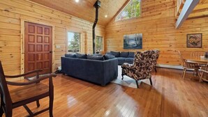 A wooden cabin interior with a sofa, armchair, dining table, and a large window. - Enjoyable 4-bedroom cabin with AC in Niagara, on a private 56 acre lake (Niagara)