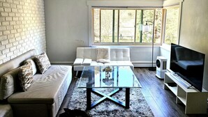 Ein Wohnzimmer mit einer Couch, einem Glastisch, einem Fernseher und einem Fenster mit Blick auf Bäume.