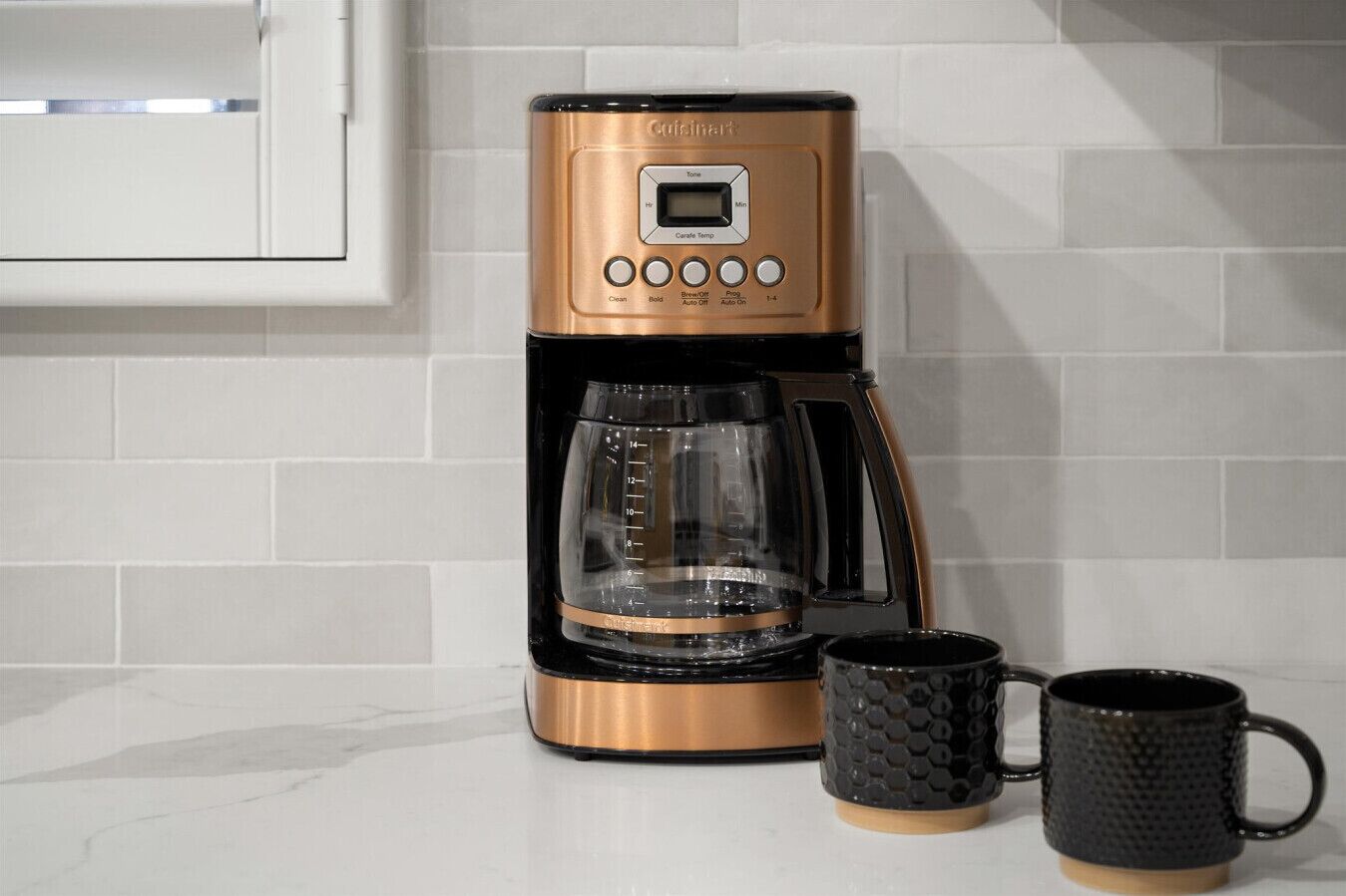Une machine à café dotée d’un écran numérique, d’une verseuse en verre et de deux tasses noires posées sur un plan de travail de cuisine.