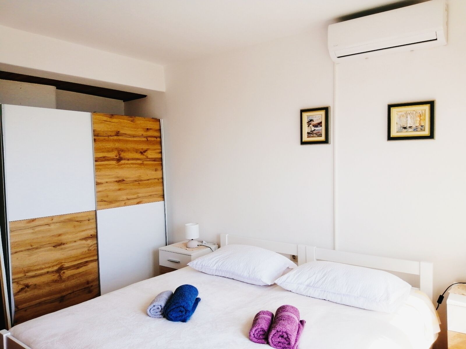 A hotel room with a bed, white pillows, and rolled towels in blue and purple. There are two framed pictures on the wall and a white nightstand with a lamp.