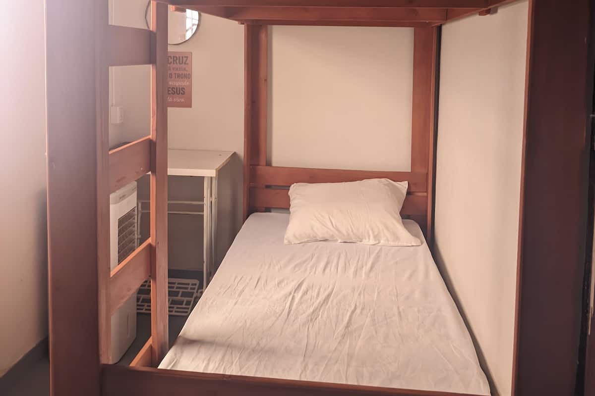 A bunk bed with a lower bunk and a single bed. There is a small shelf unit to the left and a wall-mounted heater below the lower bunk.