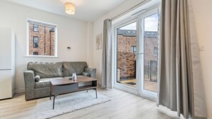 A modern living room with a grey leather sofa, a wooden coffee table, and a white rug. There is a large sliding glass door leading to an outdoor area with a brick wall and a metal fence. - Elegant Home | Parking | Wi-Fi | Near O2 Academy (Birmingham)