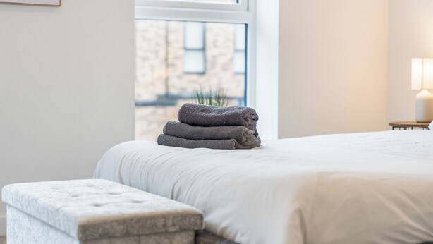 A neatly made bed with a white comforter and a stack of folded towels on the headboard. - Elegant Home | Parking | Wi-Fi | Near O2 Academy (Birmingham)