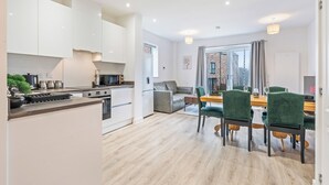 A modern kitchen with white cabinets, a dining area with a wooden table and green chairs, and a view through a glass door to the outside. - Elegant Home | Parking | Wi-Fi | Near O2 Academy (Birmingham)