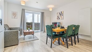 A modern dining area with a wooden table and green upholstered chairs, a grey sofa, and a large window with curtains. - Elegant Home | Parking | Wi-Fi | Near O2 Academy (Birmingham)