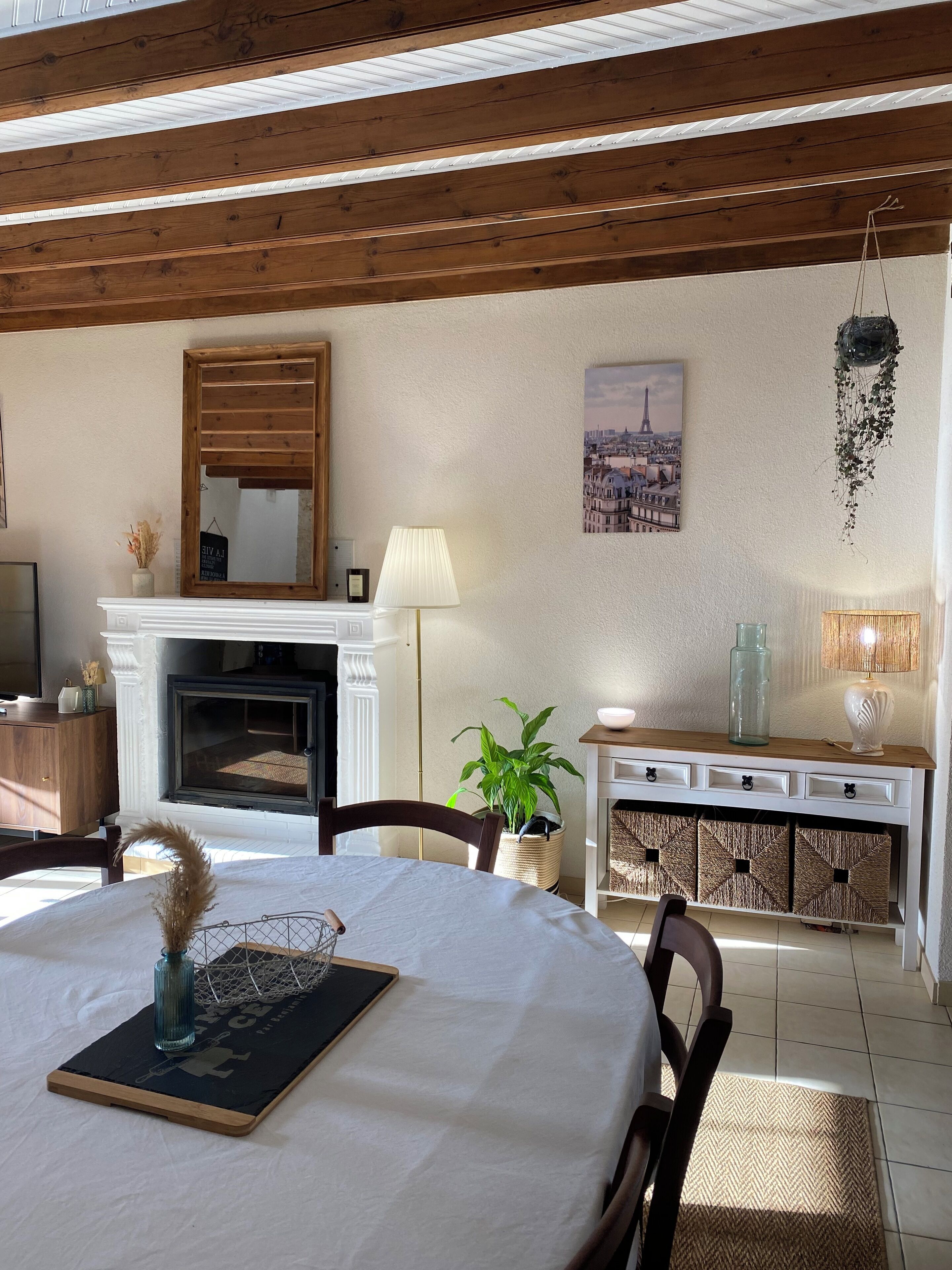 Habitació acollidora amb llar de foc, bigues de fusta, una taula de menjador amb un ram i un quadre de paisatge urbà.