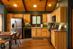 A kitchen with wooden cabinets, a stainless steel refrigerator, a dishwasher, and a dining area with a table and chairs. - Retreat•50 Acres•Mtn Views• Hot Tub•Fire Pit•Sleeps 10•Game & Media Room•Ellijay (Ellijay)