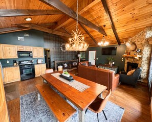 A rustic interior with wooden beams, a dining area, kitchen, and a stone fireplace. - Retreat•50 Acres•Mtn Views• Hot Tub•Fire Pit•Sleeps 10•Game Room•Carters Lake (Ellijay)